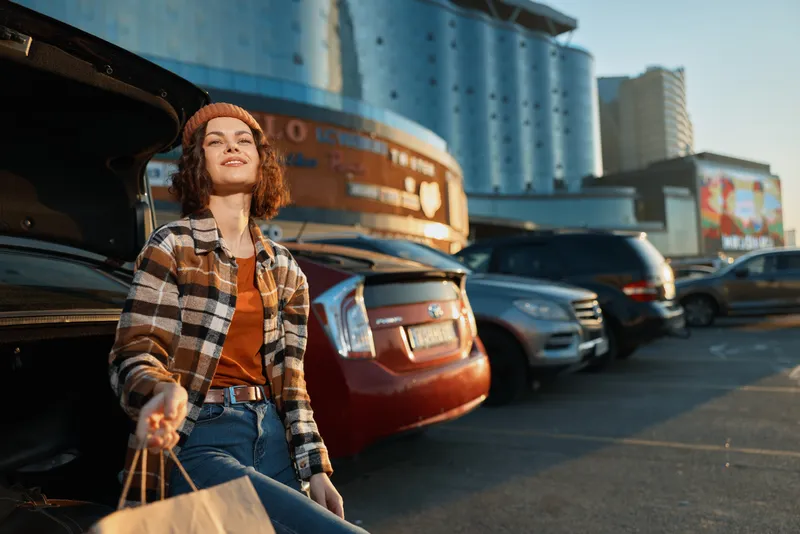 Woman shopping with bags by an open car trunk in urban parking at golden hour, candid lifestyle portrait showing authenticity, mindful living and emotional storytelling in city scene