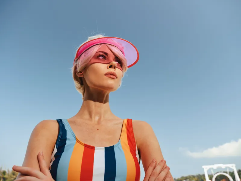 Bright portrait of a confident woman outdoors wearing a colorful striped swimsuit and pink visor, posing with crossed arms under a clear blue sky and sunlit scenery.