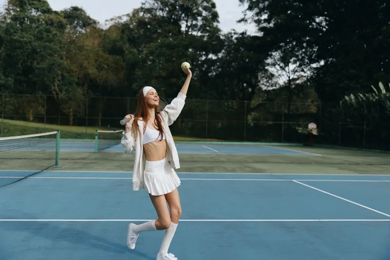 Energetic young woman in white sportswear playing tennis, joyfully serving with a tennis ball on a sunny day on an outdoor court surrounded by green trees