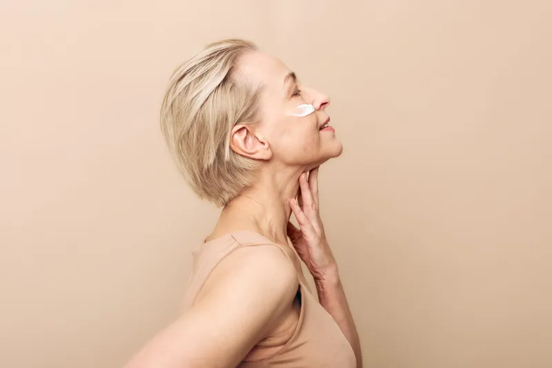 Profile of mature woman with short blond hair applying under eye patches and touching neck, smiling softly on beige background. Concept of skincare, anti aging, and self care.