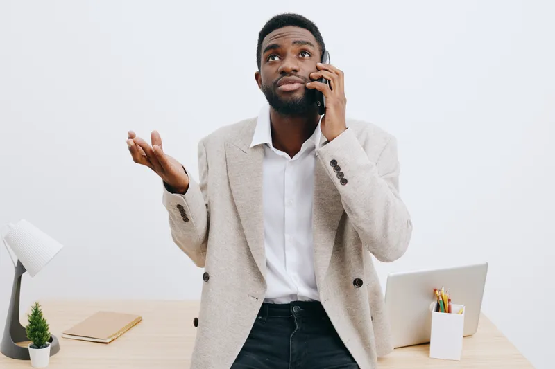 Professional young man with dark skin talking on phone while gesturing with hand, dressed in smart casual suit, standing in office with white background, studio shot, business communication concept.