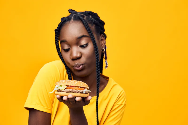 Young Black woman in a yellow shirt holds a delicious burger against a vibrant yellow background, conveying joy and satisfaction in healthy eating