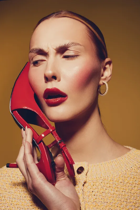 Young woman with red lipstick holds red high heel shoe near face, eyes closed and frowning. Studio portrait on yellow background. Concept of fashion, style, and beauty.