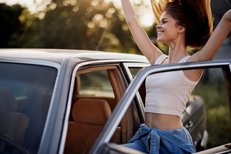Joyful young woman enjoying a road trip with her hair blowing in the wind, wearing a casual outfit in a sunny outdoor setting.