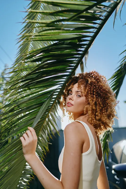 Curly woman in white tank top stands near green palm leaves under bright sunlight with a clear blue sky background, showcasing natural beauty and summer vibes.