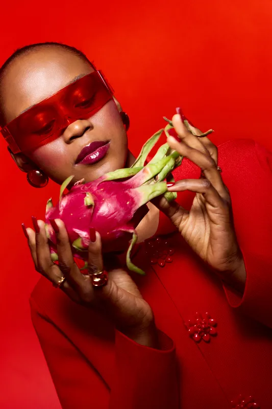 Stylish African woman with dark skin wearing red sunglasses and red outfit holds dragon fruit against red background. Concept of fashion, fruit, lifestyle, and vibrant color contrast.
