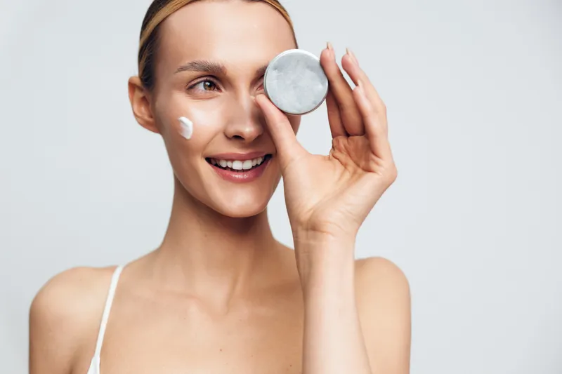 Young woman with clear skin smiles while holding a jar of cream near her eye and applying moisturizer on her cheek in a bright studio setting.