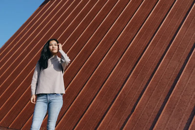 Woman standing outdoor beside a rusted metal wall, shielding eyes from bright sunlight in a casual sweater and jeans, urban contrast with diagonal lines.