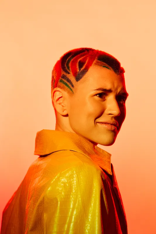 young woman with artistic red hairstyle and patterned shaved sides wearing yellow jacket, expressive facial expression, warm orange background, creative fashion portrait
