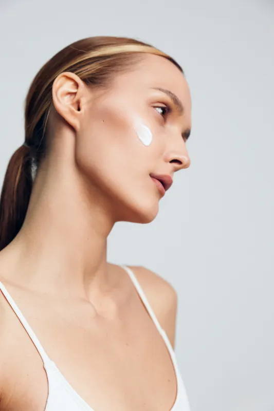Young woman with long blonde hair applies moisturizer cream on her cheek while looking sideways against a plain light background in a skincare routine moment.