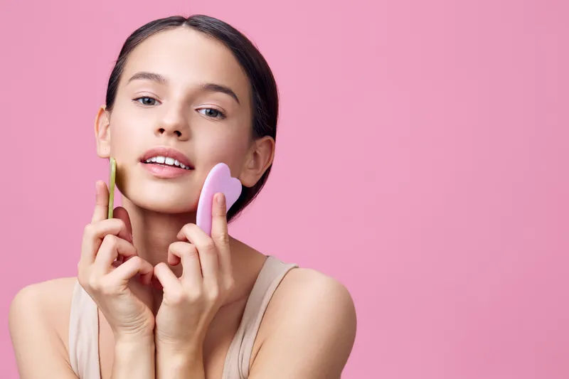 Young model using facial tools for skincare, highlighting natural beauty and wellness with pastel colors on a pink background