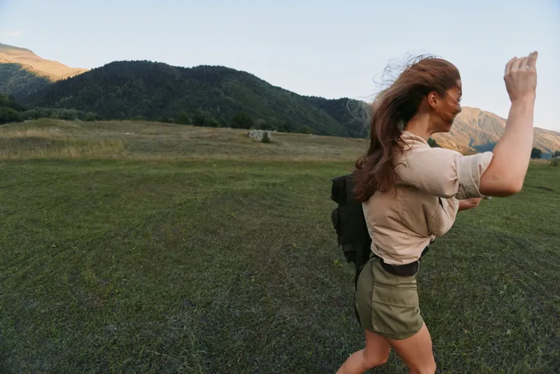 A young woman in casual clothes walks outdoors in a grassy field with mountains in the background during daylight. Concept for lifestyle, nature, and freedom.