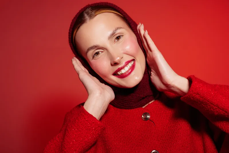 Smiling woman in red sweater and headscarf poses with hands near face against red background. Bright makeup and joyful expression create a vibrant and confident mood in studio lighting.