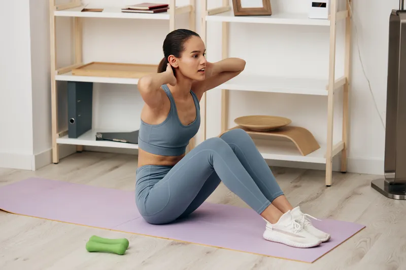 A focused athletic woman performs sit-ups on a purple mat at home, hands behind the head, blue athletic wear, with a small green dumbbell nearby and a wooden shelf in the background.