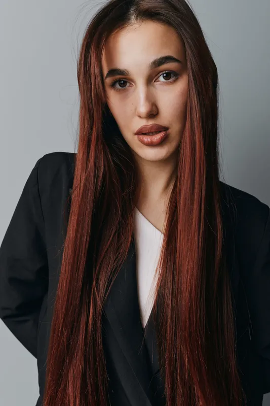 Young woman with long straight brown hair wearing black blazer and white top posing against gray background. Portrait of confident female model with natural makeup and serious expression.