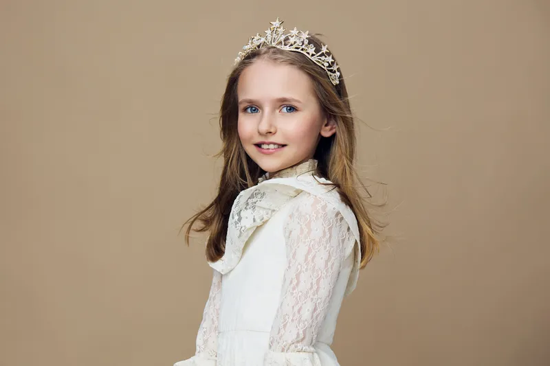 Young girl in a white lace dress and crown, smiling against a neutral beige background, reflecting innocence and joy, perfect for concepts of childhood and celebration