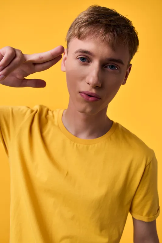 Young man in a yellow shirt makes a peace sign near his head against a bright yellow backdrop, casual fashion, confident expression, studio shot, clean composition, vibrant color mood for lifestyle