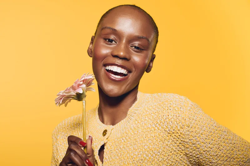 Smiling black woman with dark skin holding a pink flower against yellow background. Portrait of happy African female model wearing yellow knitted sweater. Concept of joy, beauty, and natural elegance.