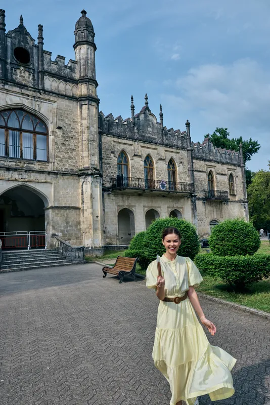 A smiling woman in a pale yellow dress walks through a castle courtyard, with ornate stone architecture and manicured hedges, showcasing timeless travel elegance under a clear blue sky.