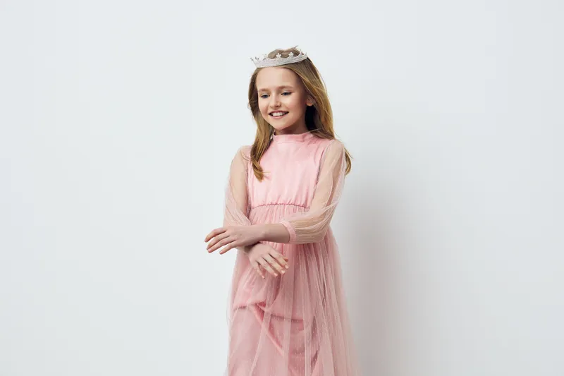 Young girl in a pink dress with a crown smiling happily, showcasing joy and innocence against a minimalistic white background, ideal for children s fashion or celebrations