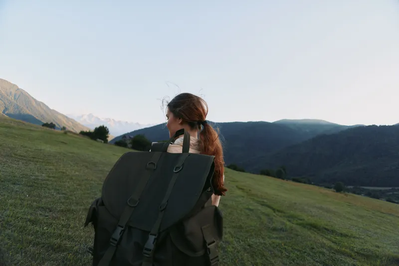 Woman hiking outdoors with large backpack, looking at mountain landscape, natural light, casual clothing, peaceful atmosphere, green grass, clear sky