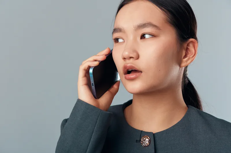 Young woman talking on smartphone wearing gray blazer with brooch on plain background. Beauty, communication, technology using mobile device for conversation and connection.