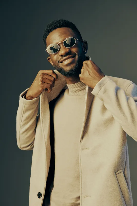 African male model in stylish sunglasses and light coat, smiling confidently against a neutral background, showcasing contemporary fashion trends