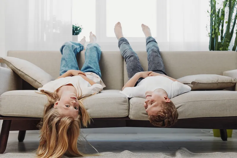 Two children lie upside down on a beige sofa in a bright living room, smiling and relaxed. Casual clothing and natural light create a cozy, playful atmosphere of childhood fun.