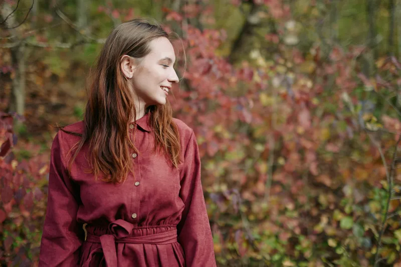 Candid outdoor portrait of a young woman in autumn foliage, smiling naturally and looking aside, conveying authenticity and credibility through genuine expression and warm seasonal tones.