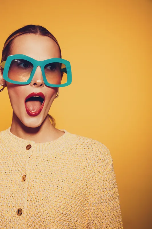 Surprised young woman with bright red lipstick wearing large turquoise sunglasses and yellow knitted sweater on yellow background. Concept of fashion, style, and vibrant emotions.