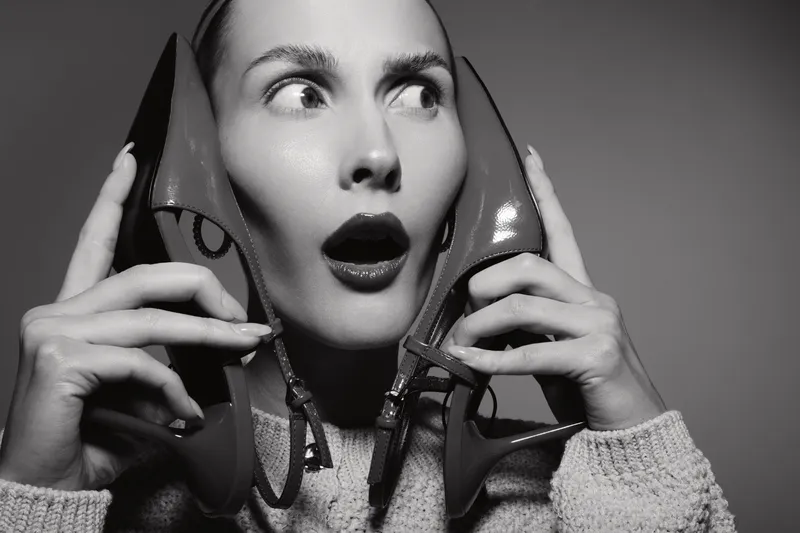 Surprised Caucasian woman with light skin holds high heel shoes near her face, wearing knitted sweater in studio lighting. Concept of fashion and emotions.