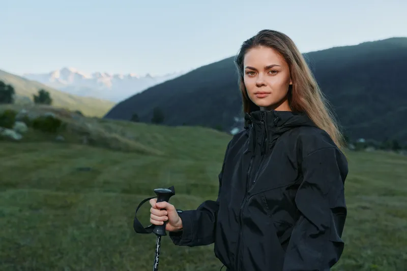 A focused adventurer stands in a green valley with a trekking pole, wearing a dark jacket. She gazes toward distant peaks, conveying determination and rugged outdoor exploration.
