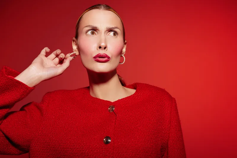 Stylish woman in red sweater poses against red background with thoughtful expression, hand near ear. Concept of fashion, beauty, portrait, and vibrant color contrast in studio lighting.