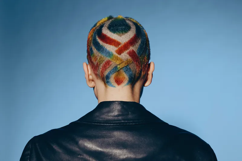 Woman with colorful geometric hair design and shaved head wearing black leather jacket against blue background, showcasing creativity and modern style.