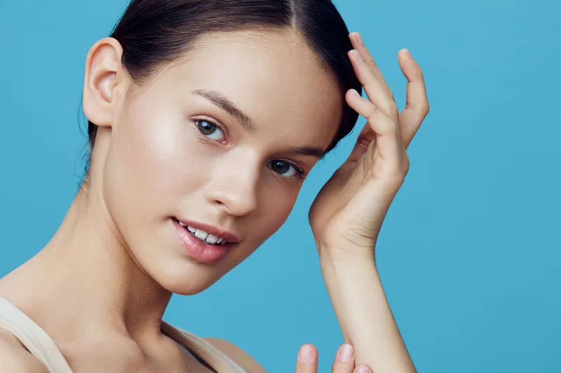 Young woman with clear skin touching her hair, against a soft blue background, representing beauty and confidence in a fresh and modern style