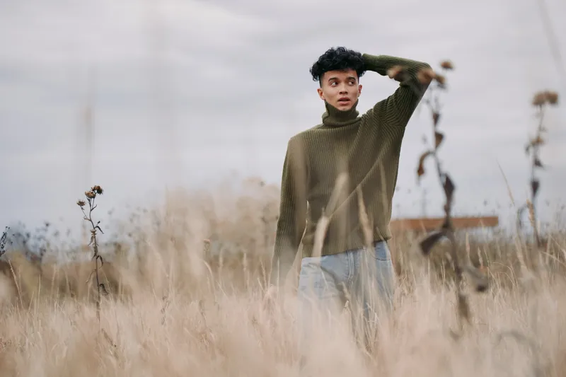 A candid portrait of a young man standing in a windswept field, captured with authenticity and credibility, highlighting natural light, textured knitwear and a thoughtful, introspective gaze.