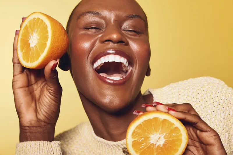 Happy dark skinned African woman laughs joyfully holding two fresh orange halves near face on yellow background. Concept of healthy lifestyle, vitamin c, nutrition and positive mood.