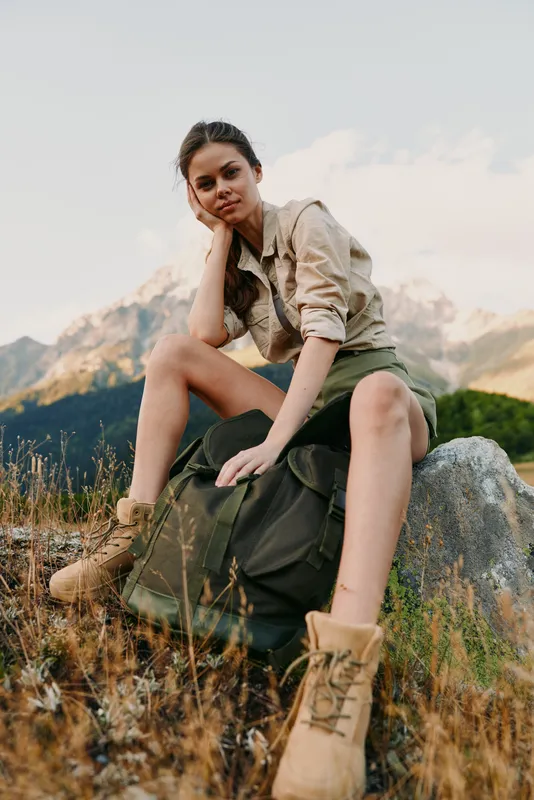 Young woman sits on a rock during a hike, wearing a backpack and rugged boots, gazing calmly at the camera with a determined adventurous vibe