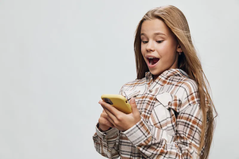 Girl with long hair looking surprised while using smartphone wearing checkered shirt. Teenager holding yellow phone with amazed expression on gray background.