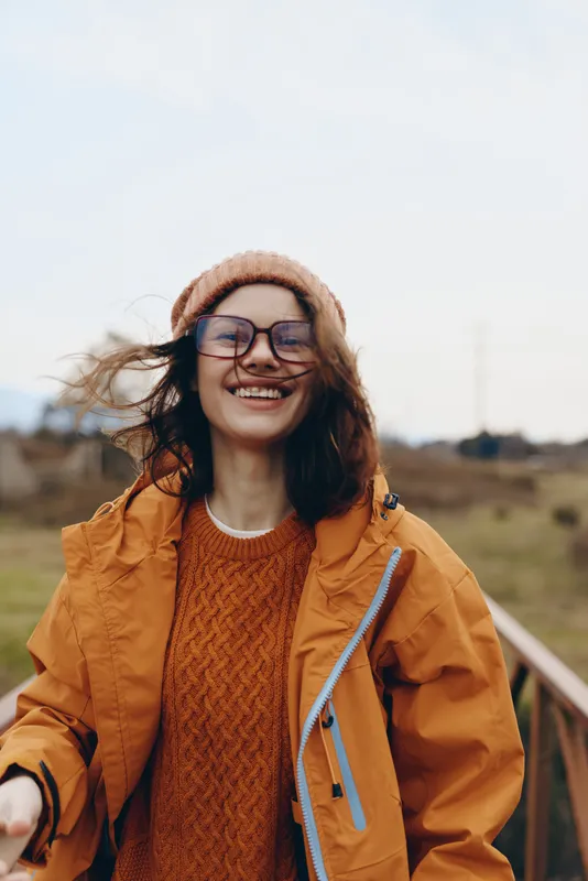 Woman lifestyle outdoor smiling in orange jacket and knit sweater, beanie hat and glasses, standing on wooden path in countryside landscape, wind moving hair, daylight warm casual portrait.