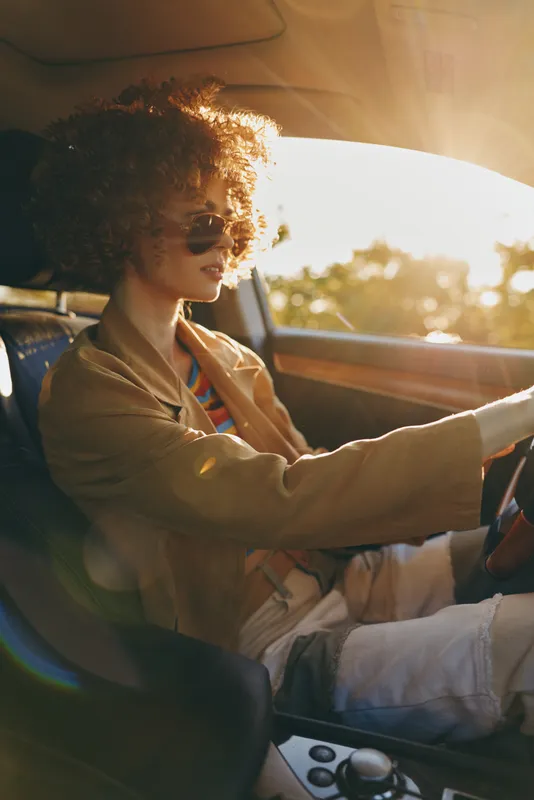 Cheerful woman driving a car, sunlit moment, casual jacket, curly hair, sunglasses, soft breeze, retro film color, natural tones, relaxed posture, pleasant road scenery behind