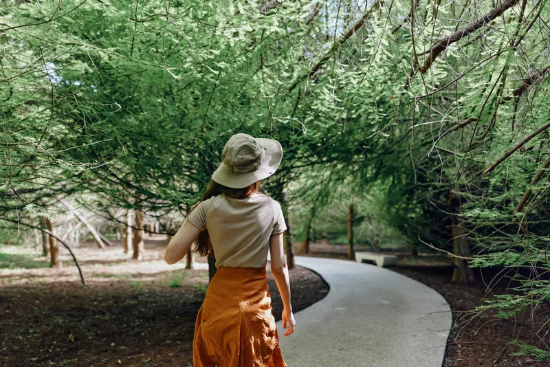Woman walking on a winding path through a green forest wearing a sun hat and orange skirt, back view. Nature trail under tree canopy, peaceful outdoor stroll and summer exploration.