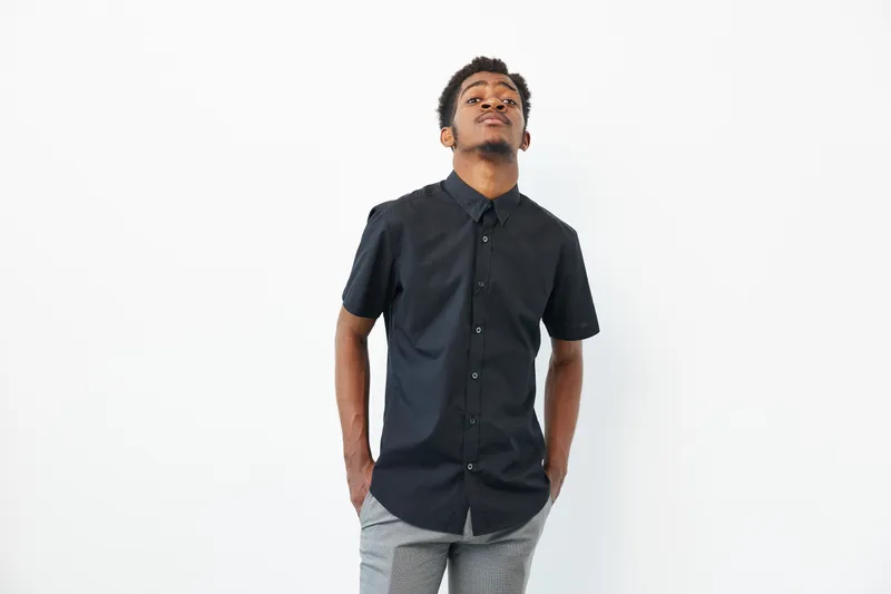 Black man wearing a stylish black shirt with confidence, expressing a thoughtful mood against a clean, minimalistic background. The image captures contemporary fashion beautifully.