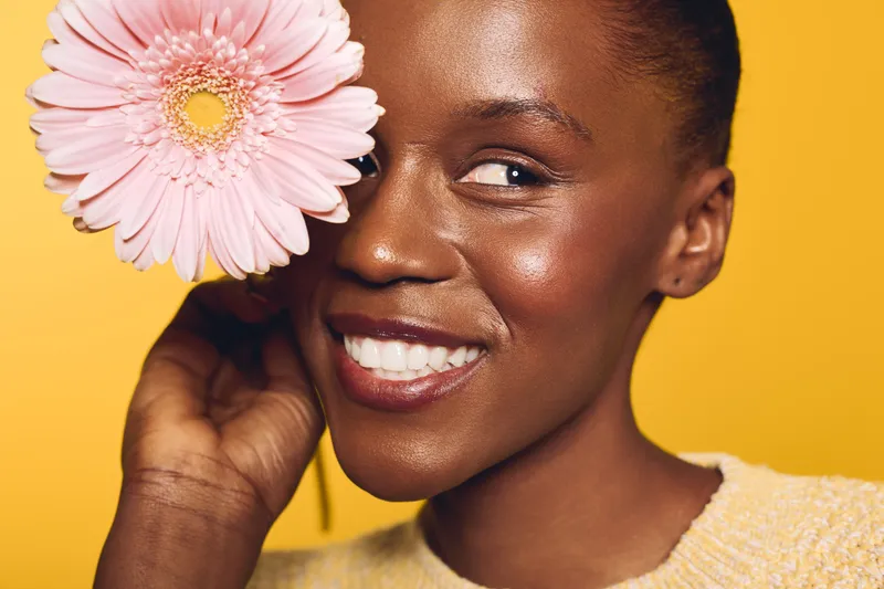 Smiling dark skinned African woman with natural makeup holds pink flower near face on yellow background. Concept of beauty, skincare, happiness, and natural portrait with floral element.