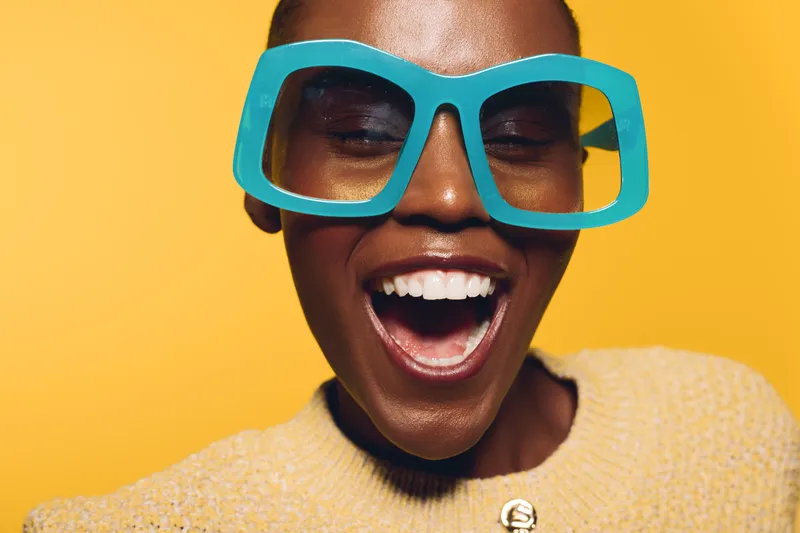 Happy African woman with dark skin wearing large blue glasses and yellow sweater smiles widely on bright yellow background. Concept of joy, fashion, and positive emotions.