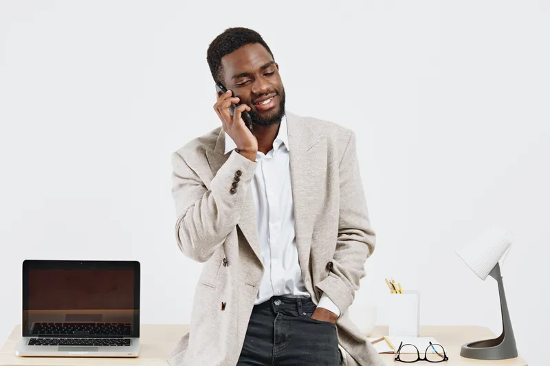 Friendly smiling man talking on the phone in modern office setting, wearing casual sweater and shirt, laptop and eyeglasses on desk, cheerful mood, professional workspace, communication concept, white