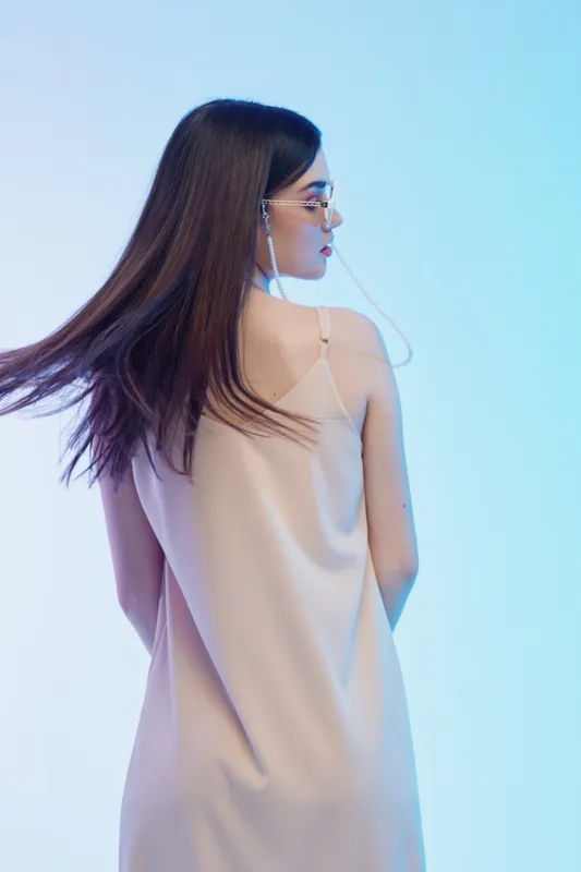 Young woman with long hair wearing glasses and a light dress poses in profile against a soft blue background. Concept of beauty, fashion, and elegance.