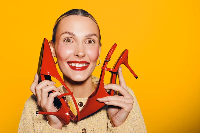 Happy woman with bright red lipstick holds two red high heel shoes near her face on yellow background. Concept of fashion, style, footwear, and joyful expression.