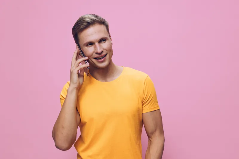 Happy young man talking on phone wearing orange t-shirt smile pink background cyber security concept mobile communication technology