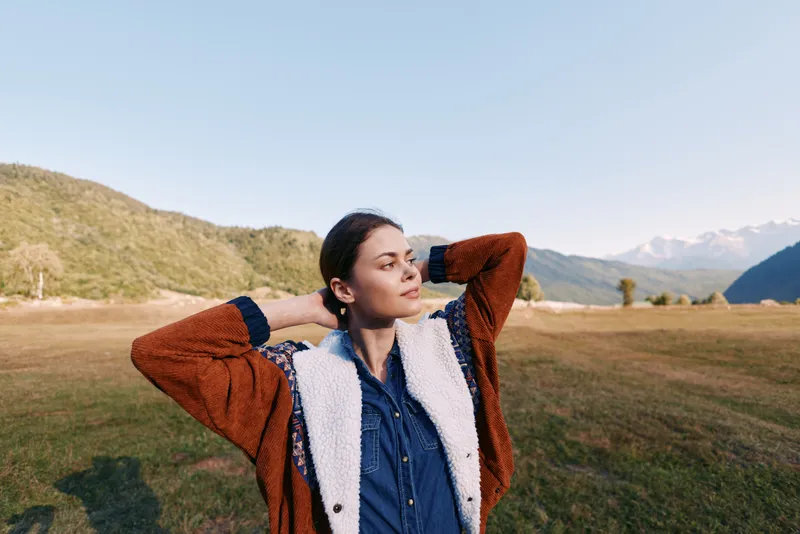 Woman portrait in mountains meadow outdoors, young relaxed in warm jacket looking into distance. Nature landscape scene with soft light, calm expression and open field.